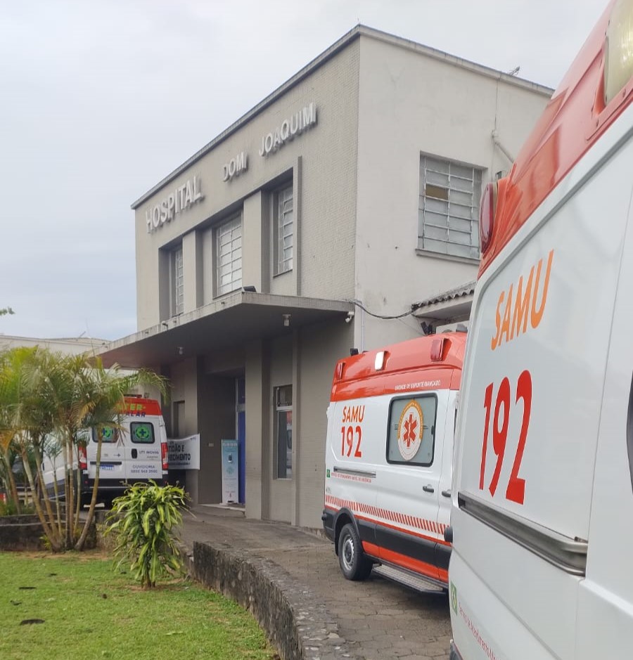 Entrada do Hospital Dom Joaquim, prédio de dois andares com fachada cinza e janelas retangulares. Em frente, estão estacionadas três ambulâncias, duas delas do SAMU com o número de emergência 192 em destaque e outra de um serviço de resgate particular. Há um pequeno jardim com plantas e gramado ao lado da calçada que leva à porta principal.
