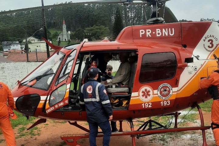Fotografia de um helicóptero de resgate vermelho e branco, identificado como "PR-BNU" e "Arcanjo 3", pousado em solo de terra. Profissionais de resgate, usando uniformes do SAMU e trajes laranja, auxiliam uma pessoa dentro da aeronave. Ao fundo, há uma área urbana com casas, árvores e uma igreja com torre alta.
