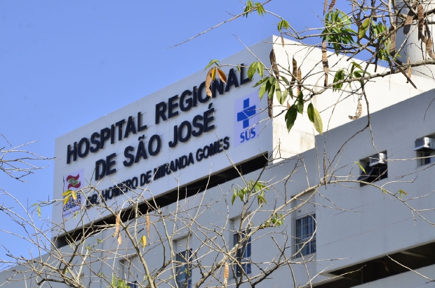 Placa na fachada do Hospital Regional de São José Dr. Homero de Miranda Gomes, com logotipos do SUS e do Governo, parcialmente encoberta por galhos de árvore.