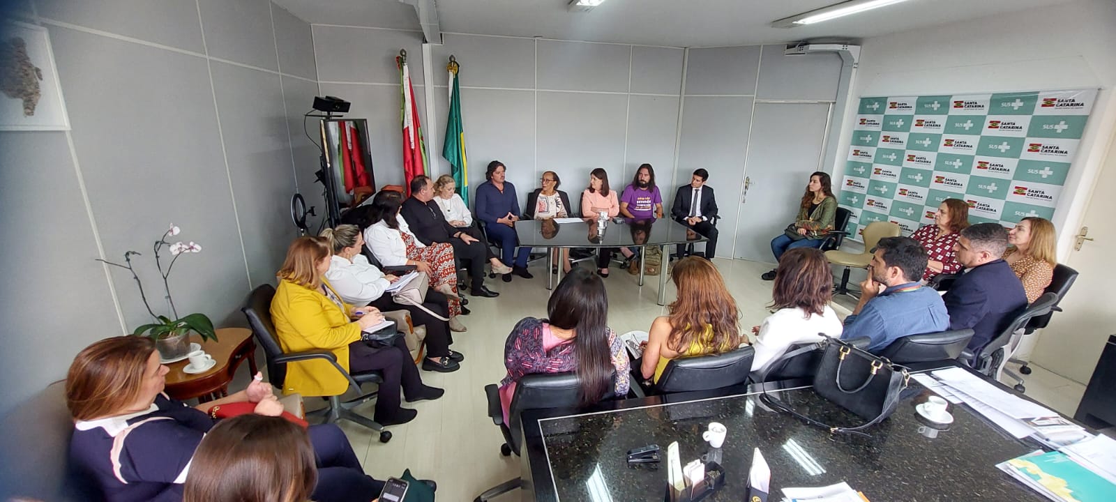 Reunião em ambiente institucional com diversas pessoas sentadas em roda, incluindo profissionais de saúde e representantes da gestão pública. Ao fundo, bandeiras do Brasil e de Santa Catarina, além de painel com logotipos da Secretaria de Estado da Saúde.