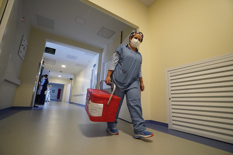 Profissional da saúde caminha por corredor hospitalar vestindo roupas cirúrgicas, touca e máscara. Ela carrega uma caixa térmica vermelha identificada como contendo um órgão para transplante. O ambiente é limpo e iluminado, com sinalizações hospitalares nas paredes.