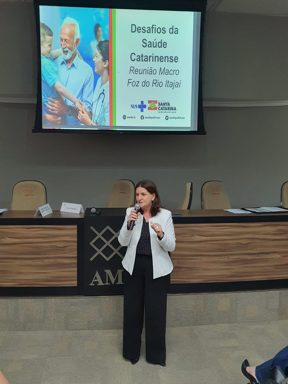 Mulher fala ao microfone em auditório durante evento institucional. Ela veste blazer branco e calça preta, e está em frente a uma mesa com cadeiras e placas de identificação. Ao fundo, há um telão com o slide do evento intitulado 'Desafios da Saúde Catarinense – Reunião Macro Foz do Rio Itajaí', com logos do SUS, Governo de Santa Catarina e Secretaria da Saúde. A imagem transmite o ambiente formal de uma reunião técnica sobre saúde pública.