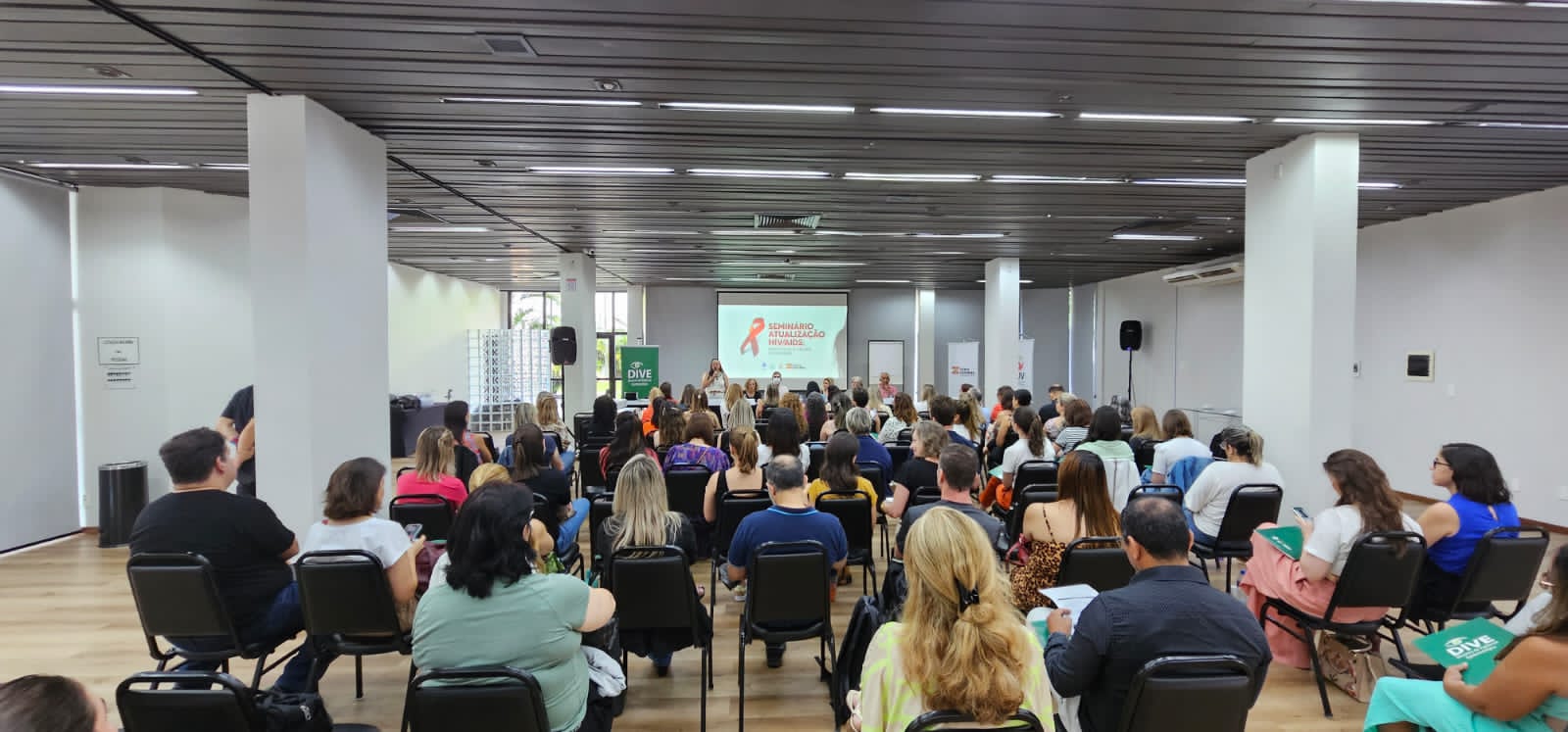 A imagem mostra um auditório cheio, com pessoas sentadas de frente para um palco onde ocorre o "Seminário de Atualização HIV/AIDS".  No telão, há um laço vermelho — símbolo internacional de conscientização sobre o HIV/AIDS — acompanhado do título do evento. À frente, várias pessoas compõem a mesa de apresentação, enquanto uma mulher em pé fala ao microfone.  Participantes, em sua maioria mulheres, estão sentados em cadeiras pretas, alguns com pastas verdes da DIVE (Diretoria de Vigilância Epidemiológica). O ambiente é iluminado, com paredes claras e teto de ripas escuras, transmitindo a atmosfera de um encontro formal e educativo.