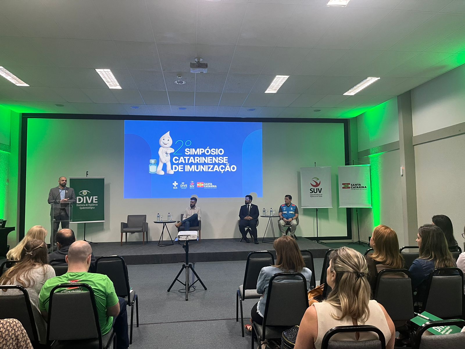 Auditório com público assistindo a uma apresentação do “2º Simpósio Catarinense de Imunização”. No palco, há um telão exibindo o logotipo do evento com a imagem do personagem Zé Gotinha. Quatro homens participam, sendo um em pé falando ao microfone e três sentados. Banners da DIVE, SUV e Governo de Santa Catarina estão posicionados ao fundo. A iluminação é predominantemente verde.'