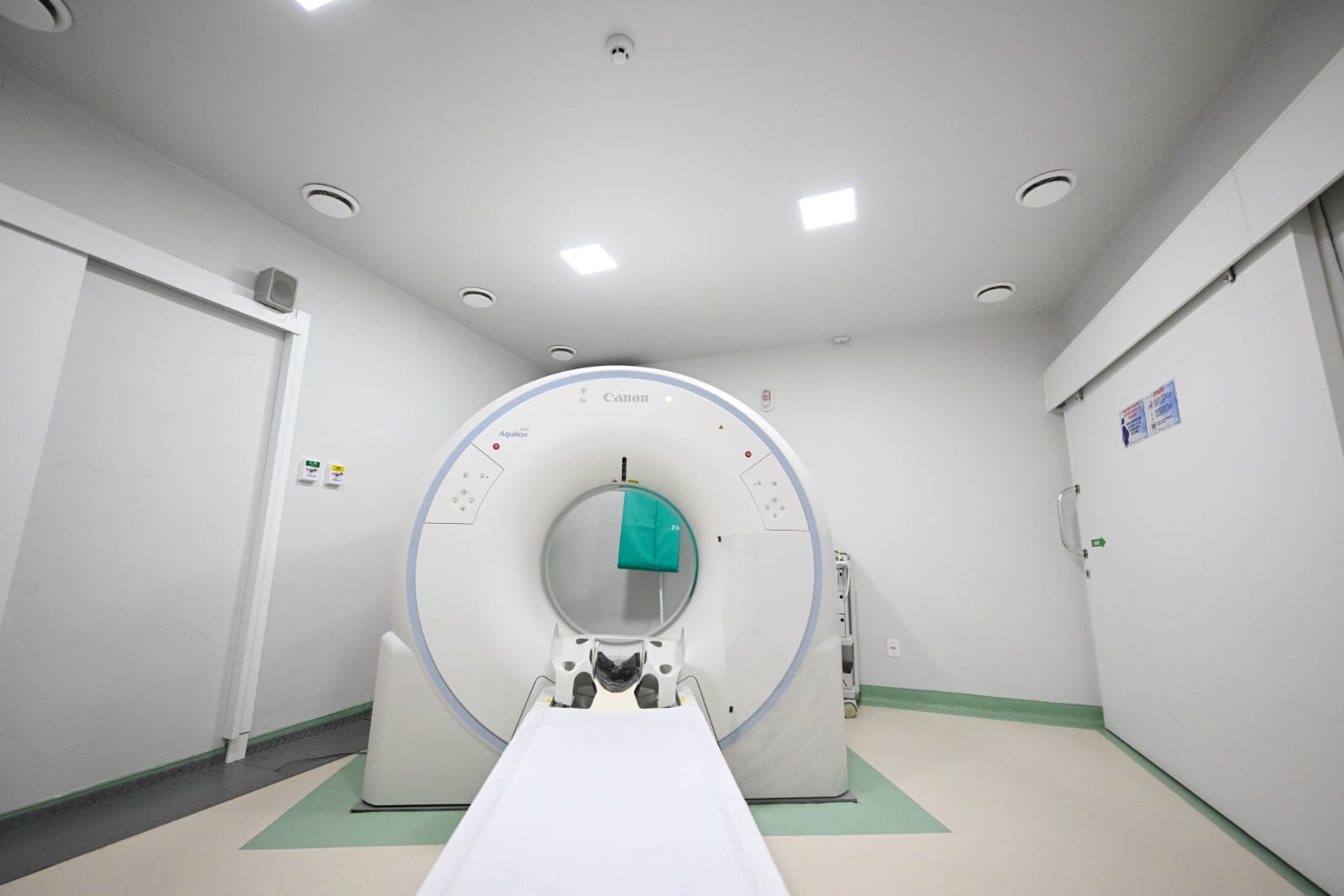 A imagem mostra um tomógrafo computadorizado instalado em uma sala hospitalar.  O equipamento é um Canon Aquilion, utilizado para exames de imagem que permitem diagnósticos detalhados do corpo humano. A sala tem paredes claras, piso em tons de verde e bege, iluminação embutida no teto e porta de correr blindada, típica de áreas com radiação ionizante.  O ambiente parece novo e bem organizado, transmitindo a sensação de que faz parte de uma estrutura recentemente inaugurada ou modernizada.