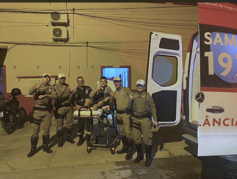 Grupo de policiais militares e profissional do SAMU ao lado de jovem em maca, posicionada próxima a ambulância com portas abertas, em ambiente externo durante a noite.