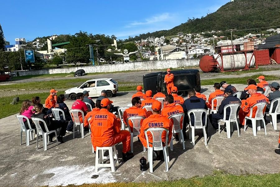 Grupo de bombeiros militares com uniformes laranja sentados em cadeiras plásticas ao ar livre, assistindo a instrução de um colega em frente a um carro capotado durante treinamento.