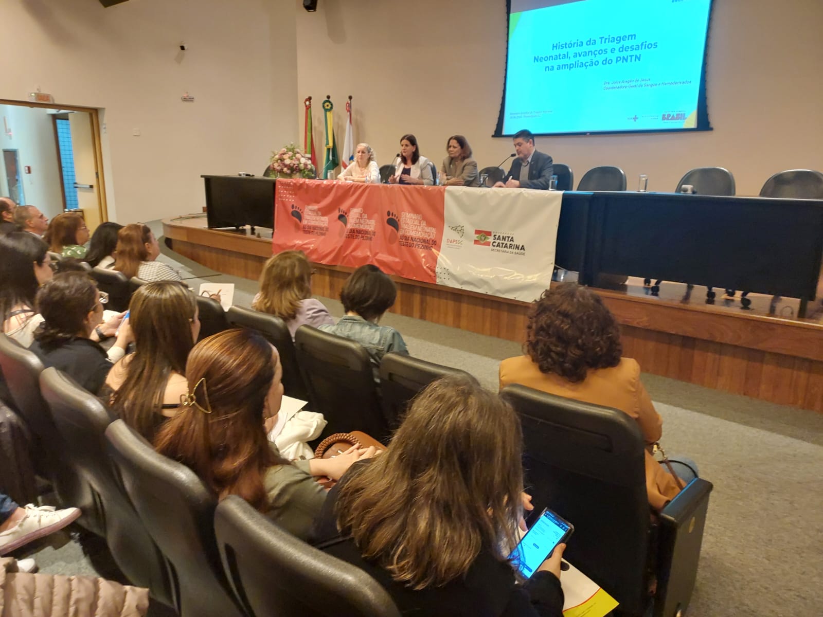 Público acompanha mesa de debate sobre a história da Triagem Neonatal em auditório, com apresentação projetada ao fundo e representantes da saúde pública de Santa Catarina.