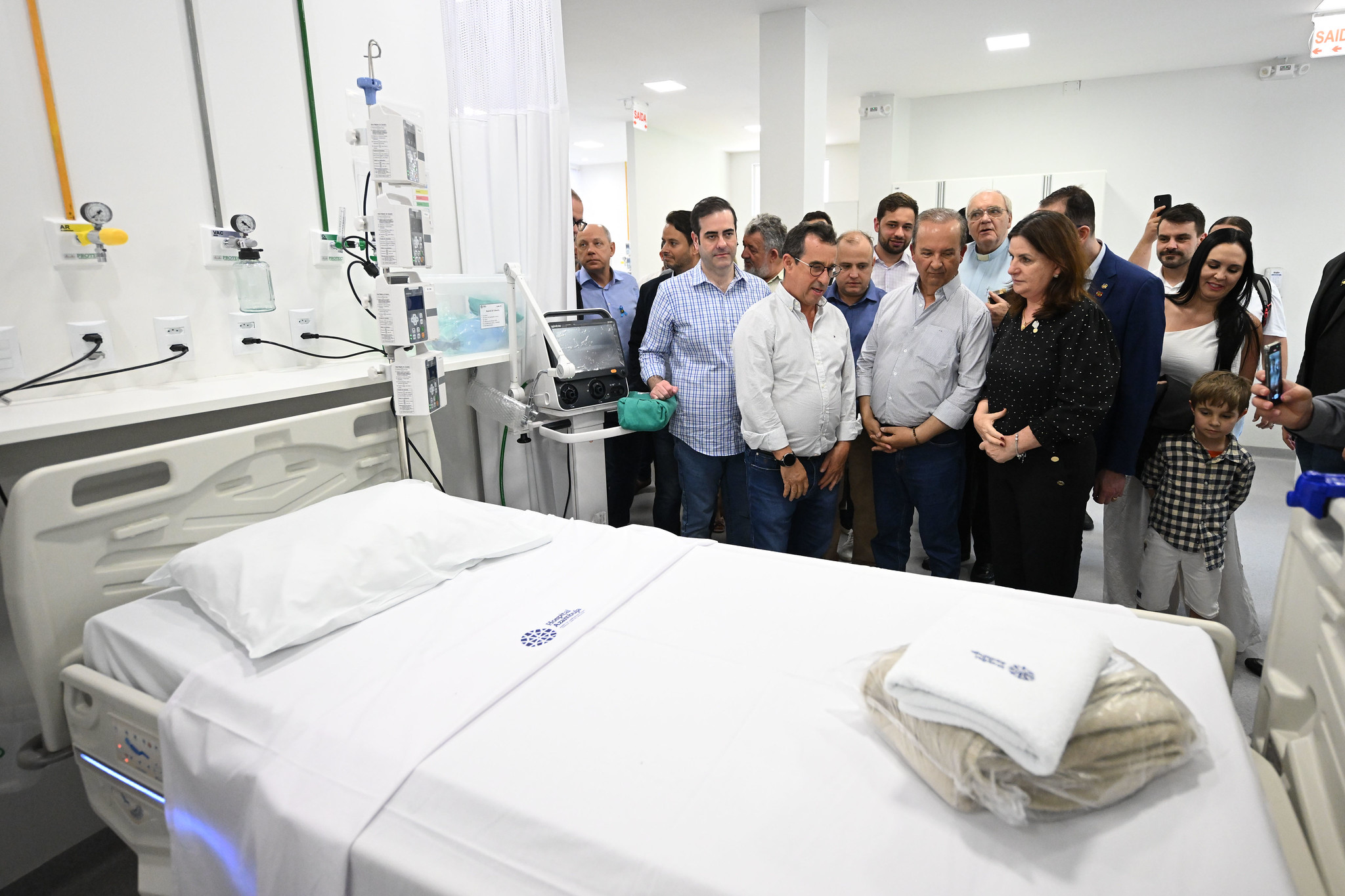 A imagem mostra um grupo de autoridades e convidados visitando uma unidade hospitalar recém-equipada, possivelmente durante uma inauguração ou apresentação oficial.  Em primeiro plano, há um leito hospitalar moderno com lençóis brancos e o logotipo da instituição, além de um cobertor e uma toalha dobrados sobre a cama. Na cabeceira, estão instalados equipamentos essenciais de UTI, como pontos de gases medicinais, monitores e bombas de infusão.  Ao fundo, várias pessoas observam atentamente, incluindo homens de camisa social e mulheres, algumas das quais parecem estar interagindo com os presentes. O ambiente é bem iluminado, limpo e organizado, transmitindo a ideia de um espaço novo e preparado para atendimento crítico.
