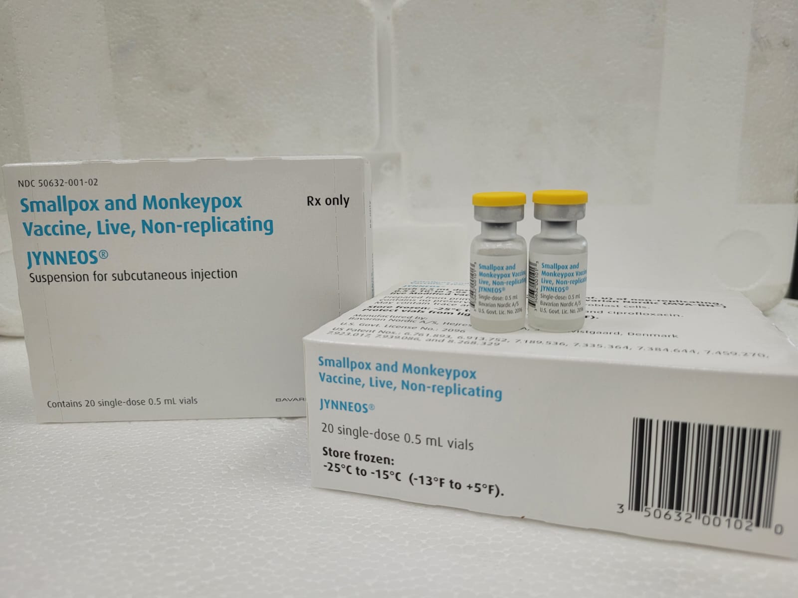 Fotografia mostra frascos e caixas da vacina JYNNEOS®, usada para a prevenção da varíola (smallpox) e da varíola dos macacos (monkeypox). Os frascos têm tampa amarela e rótulo azul. As caixas indicam que a vacina é viva, não replicante, de administração subcutânea, e deve ser armazenada congelada entre -25°C e -15°C. A embalagem contém 20 frascos de dose única de 0,5 ml.