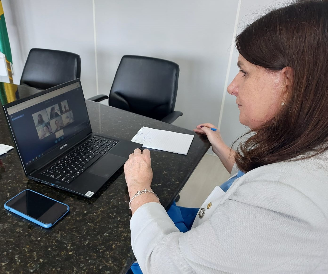 Mulher em ambiente de trabalho participa de uma videoconferência pelo notebook. Ela está sentada à mesa com um caderno de anotações e um celular ao lado. Na tela do computador, várias pessoas estão visíveis em uma chamada de vídeo. Ao fundo, duas cadeiras vazias e uma bandeira indicam um ambiente institucional. 