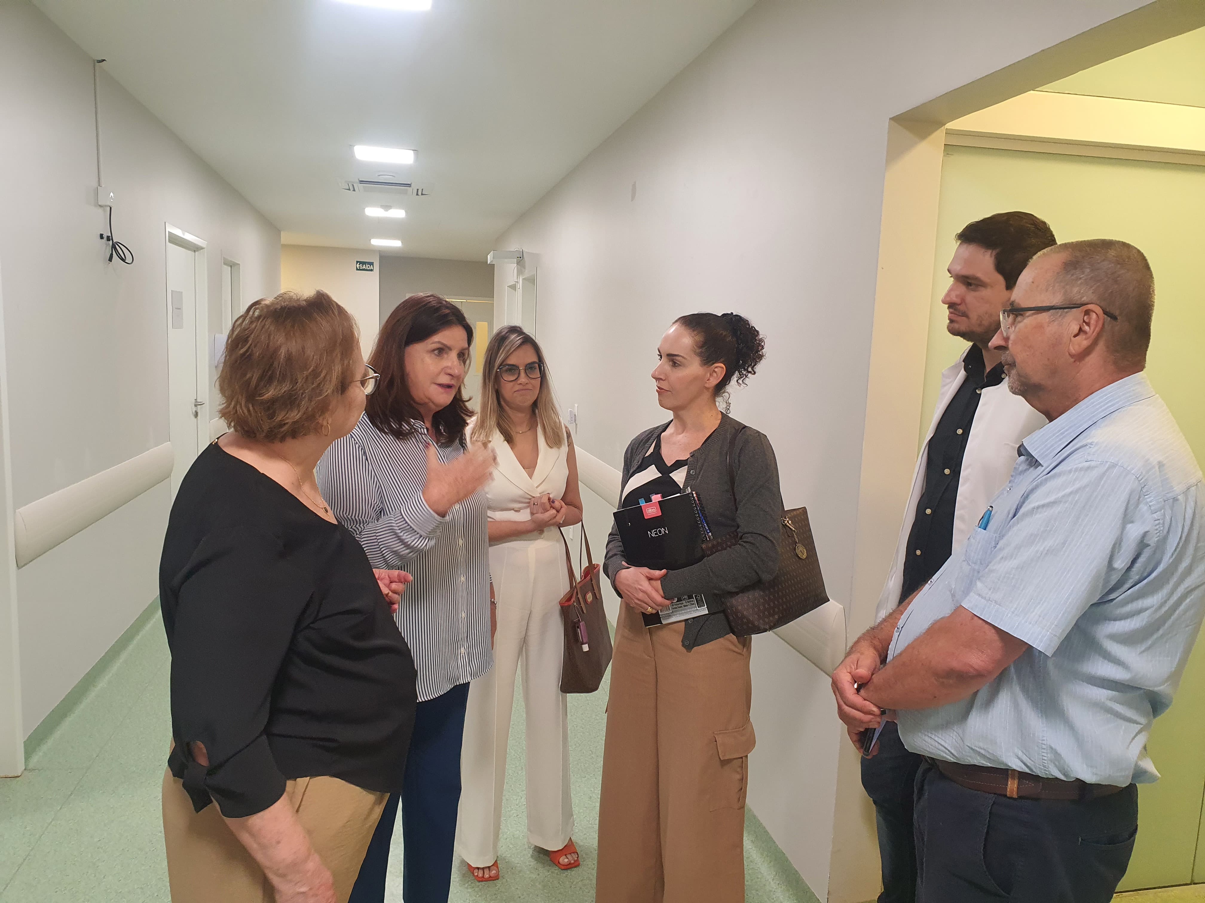 A imagem mostra seis pessoas em um corredor interno de um hospital, conversando em grupo. Ao centro, uma mulher de cabelos castanhos, usando camisa listrada em branco e azul, gesticula enquanto fala. Ao redor dela, três mulheres e dois homens escutam atentamente; uma das mulheres segura uma bolsa e um caderno, enquanto um dos homens veste jaleco branco, indicando ser um profissional de saúde. O ambiente é iluminado por luzes de teto e possui piso verde-claro, paredes brancas e portas hospitalares, reforçando o contexto de visita ou inspeção técnica em unidade de saúde.
