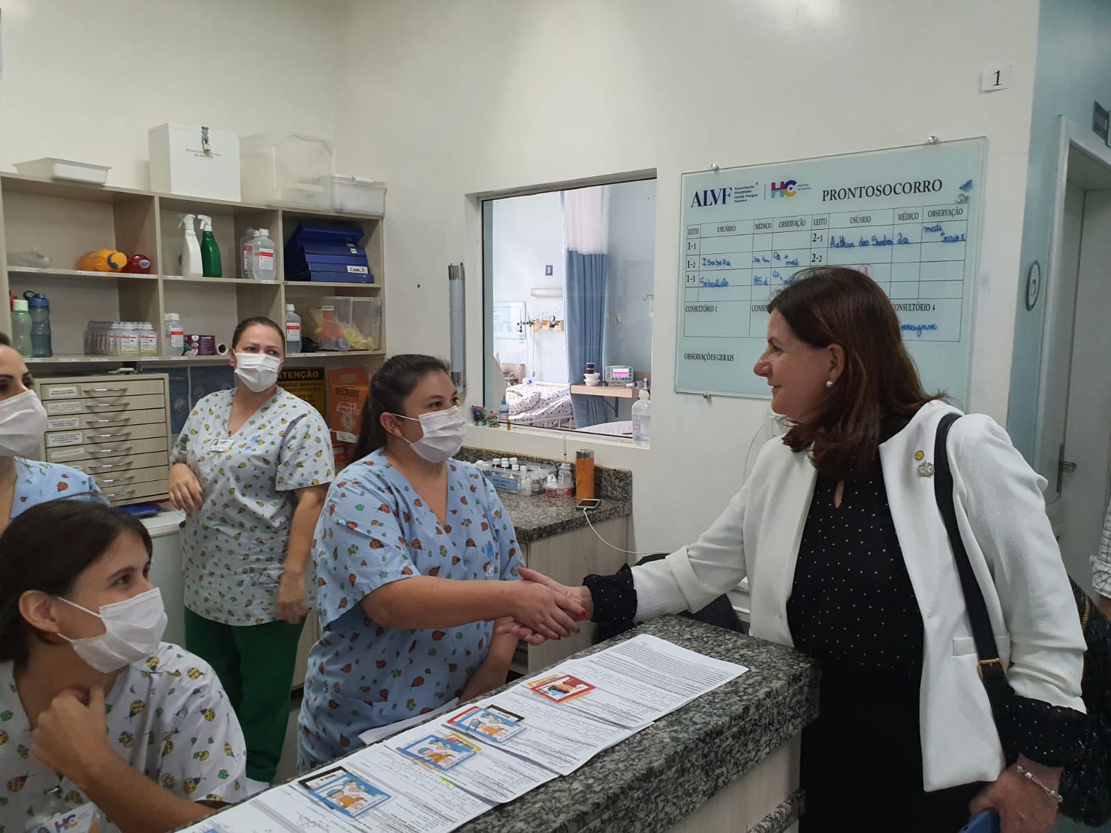 Mulher com blazer branco e blusa preta com bolinhas, usando máscara facial, cumprimenta uma profissional de saúde em um balcão de recepção hospitalar. Outras quatro profissionais, também de máscara e com uniformes hospitalares estampados, observam a interação. Ao fundo, há uma lousa branca com informações do pronto-socorro e prateleiras com materiais hospitalares.