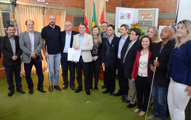 Grupo de cerca de 17 pessoas posa sorridente para foto em ambiente interno, com paredes de tijolos aparentes e cortinas claras ao fundo. No centro, um homem de blazer claro segura um documento ao lado de outras autoridades.