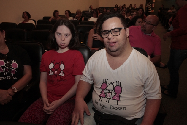 Jovem homem e jovem mulher com síndrome de Down, usando camisetas da campanha 'Amigo Down', sentados em uma plateia de auditório durante evento da Semana Inclusiva em Florianópolis, que debate a inclusão e o assédio moral no ambiente de trabalho.