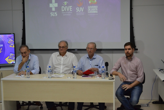 Quatro homens sentados à mesa, com projeção ao fundo exibindo os logotipos do SUS, DIVE, SUV e Governo de Santa Catarina.