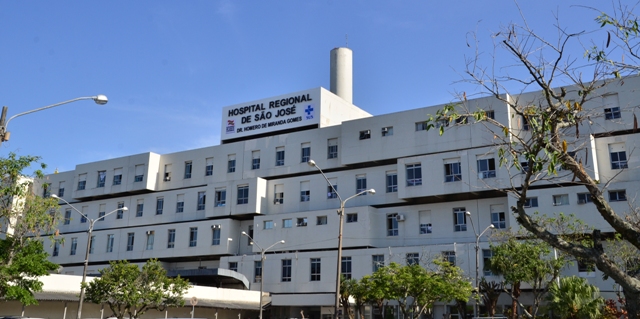 Fachada do Hospital Regional de São José Dr. Homero de Miranda Gomes, em Santa Catarina, com prédio de múltiplos andares, janelas alinhadas e nome da instituição em destaque no topo. Céu azul e árvores ao redor complementam a paisagem externa da unidade de saúde.