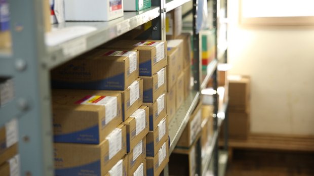 A imagem mostra um estoque organizado com caixas de medicamentos ou suprimentos médicos dispostas em prateleiras metálicas. As caixas são etiquetadas, indicando um ambiente hospitalar, farmacêutico ou de armazenamento de insumos de saúde. A luz natural entra por uma janela ao fundo, e o local parece limpo e bem iluminado. Esse tipo de armazenamento é típico de unidades de saúde ou centrais de distribuição de medicamentos.