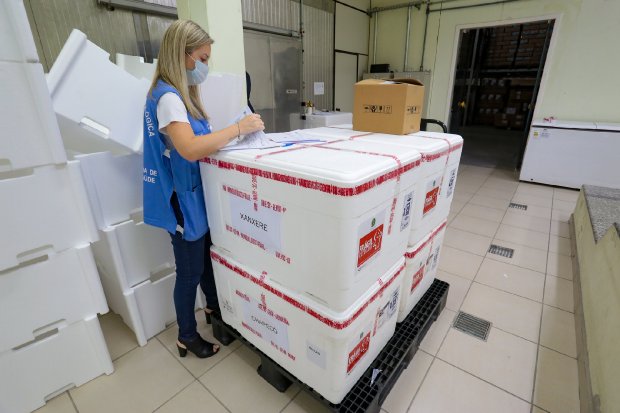 Sala com mulher loira de colete azul abrindo caixa térmica branca empilhada em outras caixas