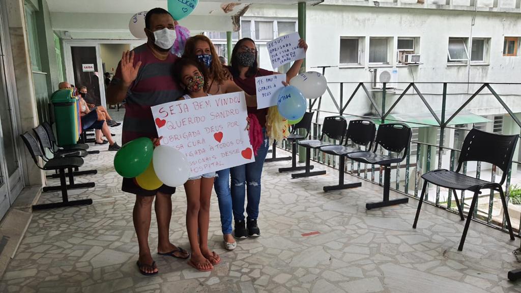 Grupo de quatro pessoas com máscaras de proteção segurando cartazes e balões coloridos em uma área externa de hospital. Um dos cartazes diz Seja bem-vindo querido Sandro. Obrigada Deus por essa cura. Obrigada a todos os profissionais desse hospital. Todos sorriem e acenam, demonstrando alegria e gratidão. Ao fundo, outras pessoas estão sentadas em cadeiras plásticas, próximas à entrada do prédio.