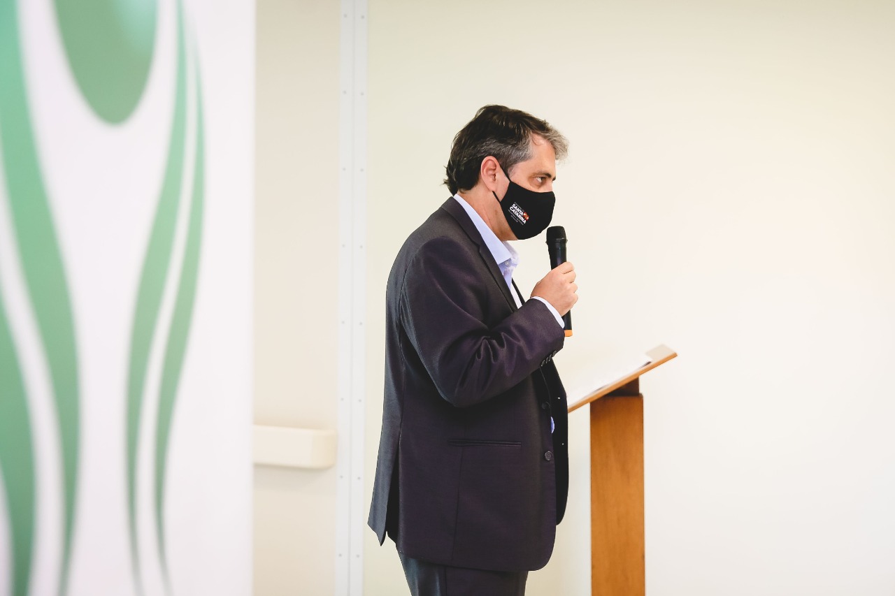 Homem de terno escuro e máscara preta com o logotipo do Governo de Santa Catarina fala ao microfone em um púlpito de madeira, em ambiente interno de paredes claras.