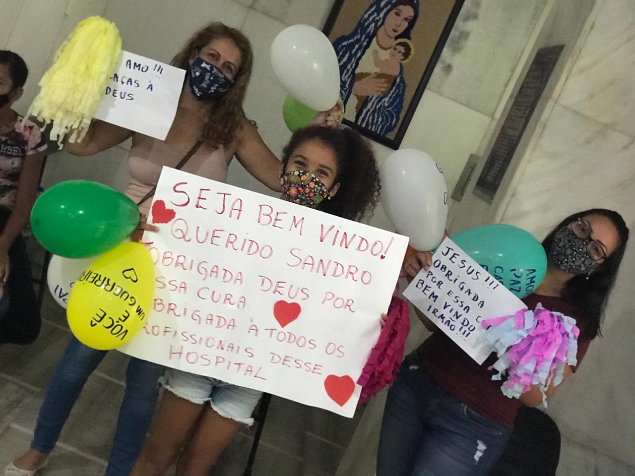Três mulheres e uma criança, todas usando máscaras, seguram cartazes, balões e pompons coloridos em um corredor interno de hospital. O cartaz principal diz: Seja bem-vindo! Querido Sandro. Obrigada Deus por essa cura. Obrigada a todos os profissionais desse hospital. Os balões contêm mensagens como Você vai conseguir e Amor, paz e cura. Ao fundo, há uma imagem de Nossa Senhora com o Menino Jesus em mosaico na parede.