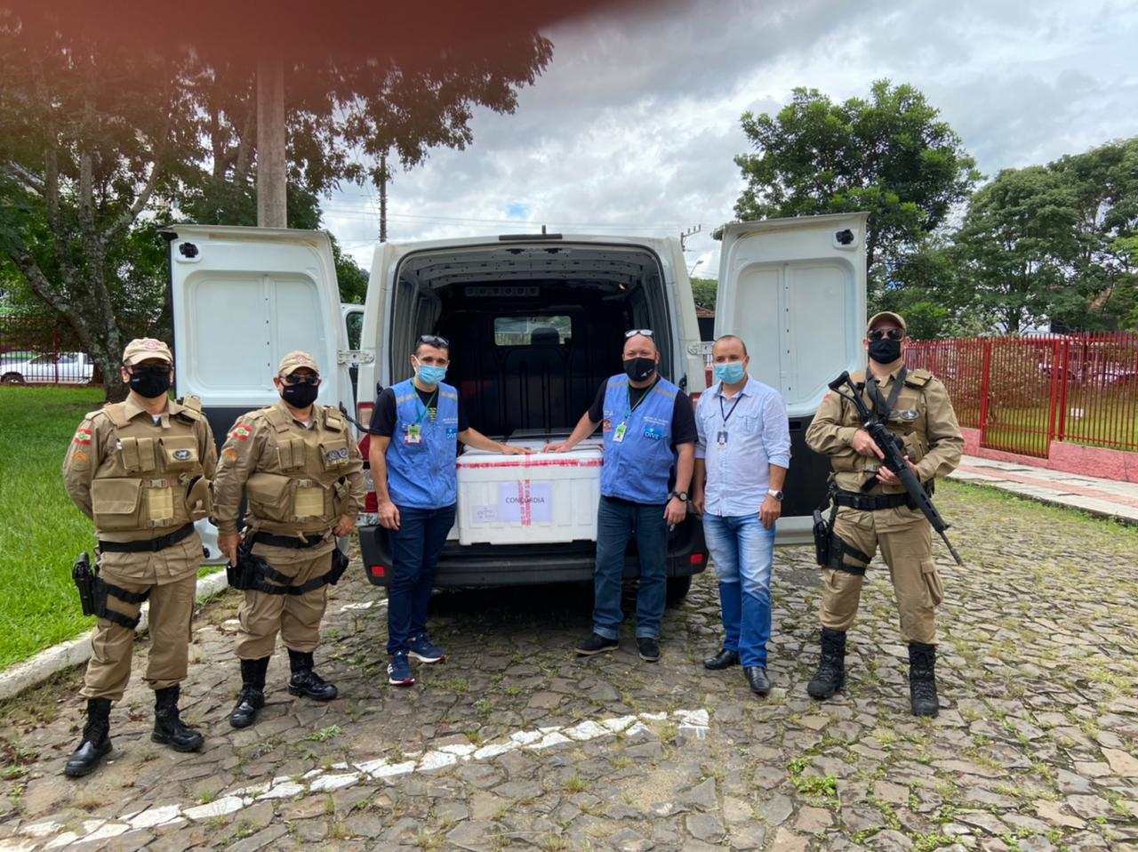 Profissionais da saúde e policiais militares posam ao lado de uma van branca com as portas traseiras abertas. Dentro do veículo há uma caixa térmica identificada como Concórdia, possivelmente contendo vacinas. O grupo usa máscaras de proteção facial e está em um pátio com calçamento de pedras, com árvores e um portão vermelho ao fundo.