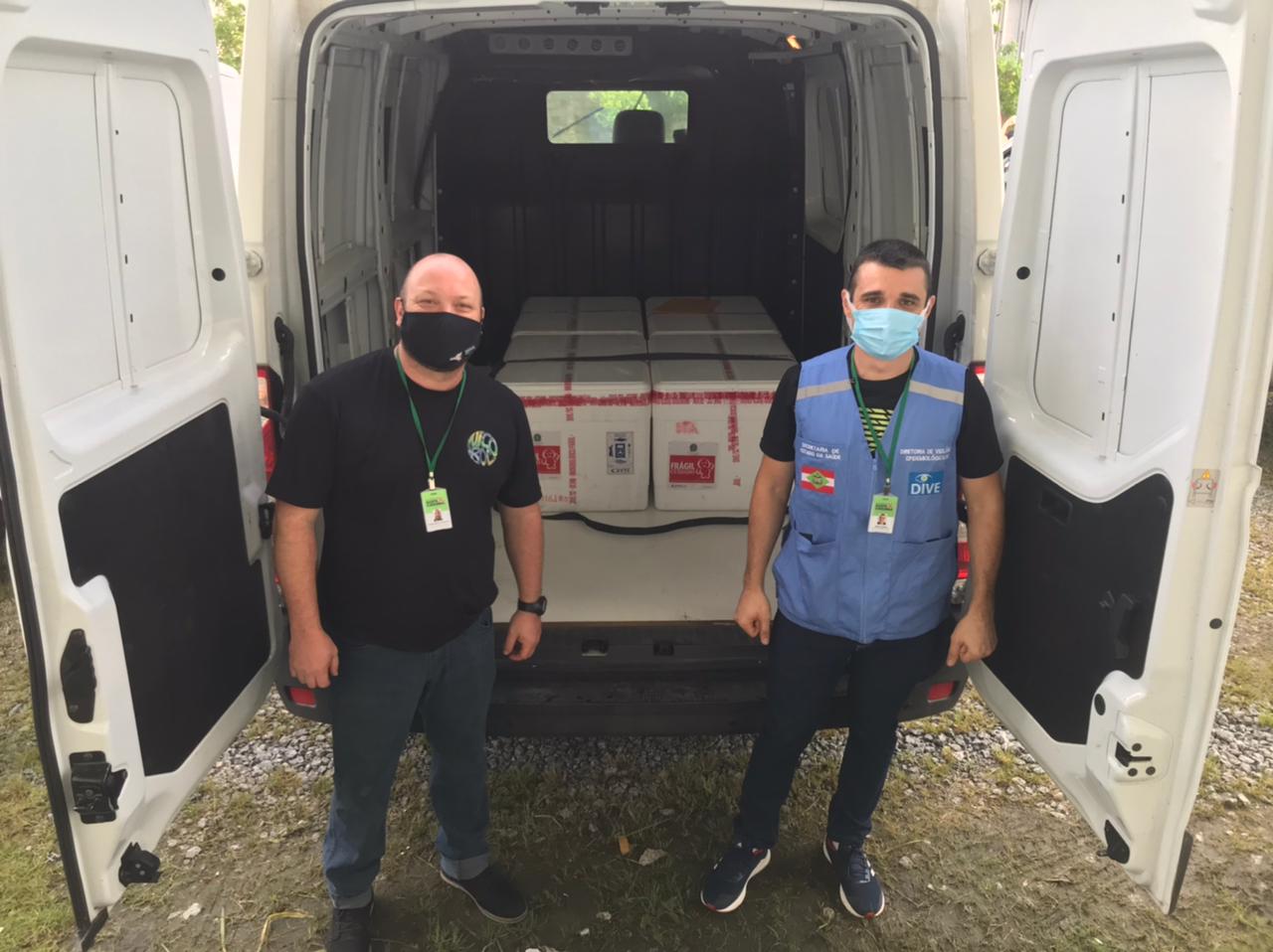 Dois profissionais da saúde posam ao lado de uma van com as portas traseiras abertas, mostrando caixas térmicas identificadas com rótulos de vacinas. Ambos usam máscaras e crachás, sendo um deles com colete da Diretoria de Vigilância Epidemiológica de Santa Catarina. A cena acontece em área externa com gramado e pedras no chão.