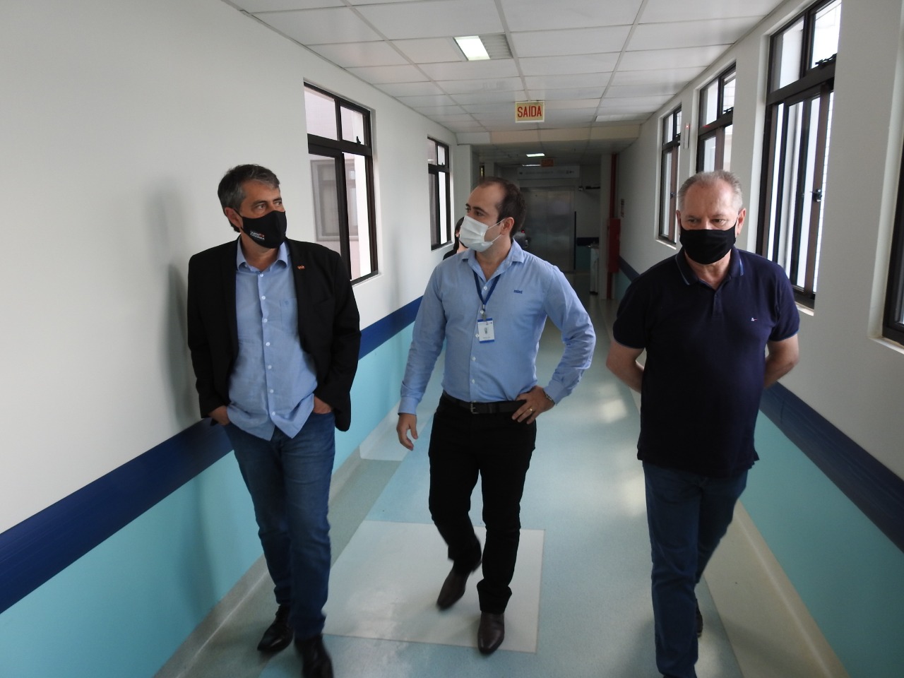 Tres homens usando máscaras caminham por um corredor de hospital iluminado, com janelas à direita e sinalização de saída ao fundo. O homem à esquerda veste paletó preto e calça jeans; o do centro está de camisa azul clara e crachá; o da direita veste camisa polo azul escuro.