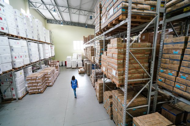 Pessoa caminha por um amplo galpão logístico com pé-direito alto, iluminado por luz natural vinda de uma janela ao fundo. O espaço está organizado com prateleiras metálicas altas, repletas de caixas de papelão e isopores empilhados, indicando armazenamento de materiais, possivelmente insumos hospitalares ou vacinas.