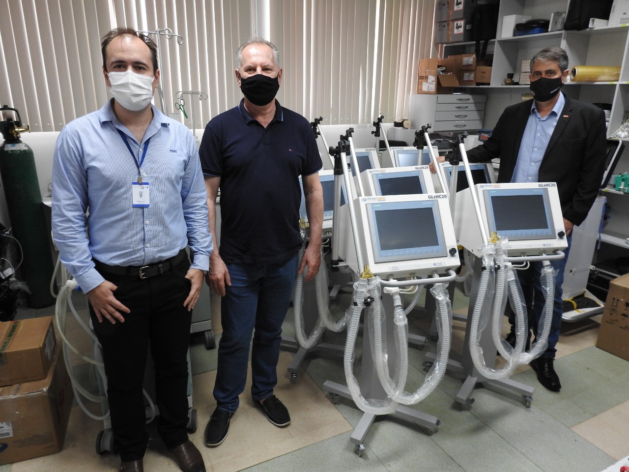 Três homens usando máscaras posam em frente a um grupo de ventiladores pulmonares hospitalares em uma sala com equipamentos médicos e caixas. O homem à esquerda usa camisa azul clara com crachá, o do centro veste camisa polo azul escuro e o da direita está de paletó preto e camisa azul.