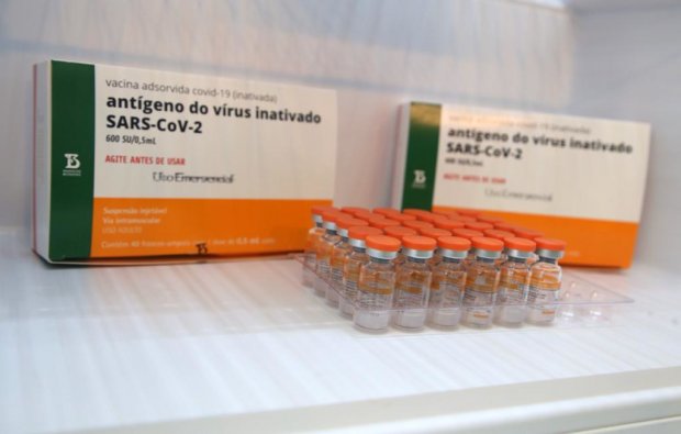 Frascos da vacina CoronaVac contra a COVID-19, dispostos sobre uma superfície branca. Ao fundo, caixas identificadas com o rótulo “vacina adsorvida covid-19 (inativada) – antígeno do vírus inativado SARS-CoV-2”, com logotipo do Instituto Butantan e menção ao uso emergencial. As tampas dos frascos são laranja e as caixas têm as cores branca, verde e laranja.