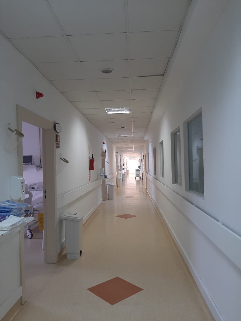 Corredor longo e bem iluminado de um hospital, com piso claro e detalhes geométricos alaranjados. As paredes brancas possuem corrimãos, janelas internas à direita e portas de acesso a quartos à esquerda. Um quarto visível à esquerda mostra parcialmente uma cama hospitalar. Equipamentos médicos e lixeiras estão distribuídos ao longo do corredor.