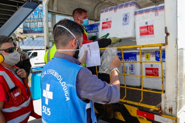Um profissional da Vigilância Epidemiológica, identificado pelo colete azul, verifica documentos enquanto outra pessoa retira uma caixa térmica branca da caçamba de um veículo. 