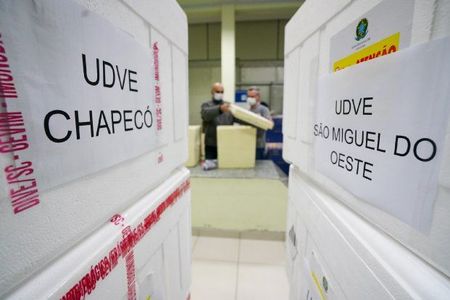Caixas térmicas brancas com etiquetas escritas UDVE Chapecó e UDVE São Miguel do Oeste em destaque. Ao fundo, duas pessoas usando máscaras manuseiam outra caixa em um ambiente interno, provavelmente relacionado à logística de vacinas ou insumos de saúde.
