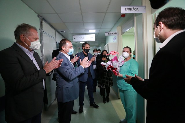 Diversas pessoas aplaudindo dentro de hospital uma mulher com roupas hospitalares verdes  segurando uma flor que ganhou de presente
