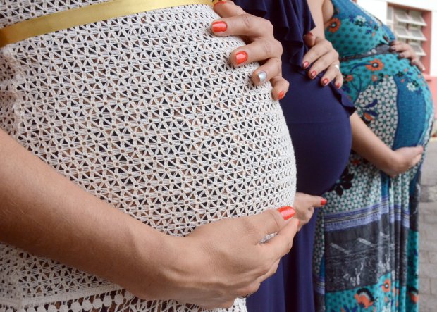 Três mulheres grávidas estão posicionadas lado a lado, com as mãos apoiadas sobre suas barrigas. A imagem mostra apenas os troncos e barrigas, destacando as diferentes roupas: uma veste um vestido branco rendado com faixa dourada, outra usa vestido azul escuro, e a terceira está com um vestido estampado em tons de azul e verde. A foto enfatiza o cuidado e a proteção à gestação.