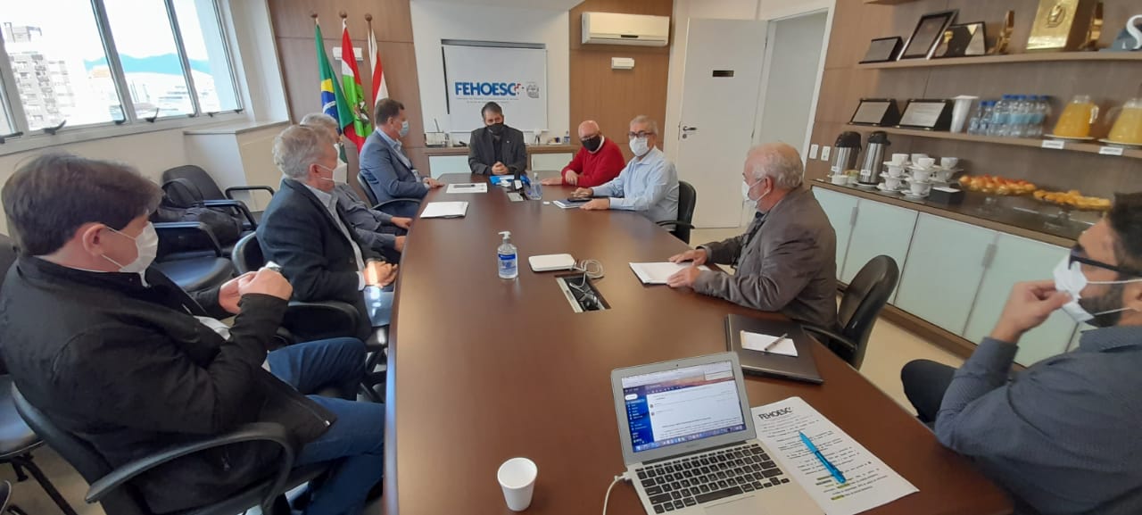 Reunião formal com nove homens sentados ao redor de uma mesa de reunião retangular na sede da FEHOESC (Federação dos Hospitais de Santa Catarina). Todos usam máscaras faciais. A mesa contém laptops, papéis, copos e álcool em gel. Ao fundo, há bandeiras do Brasil, de Santa Catarina e outra institucional. Estantes com café, sucos e petiscos estão posicionadas na lateral direita da sala, indicando um ambiente corporativo. A janela ao fundo mostra parte da vista urbana.