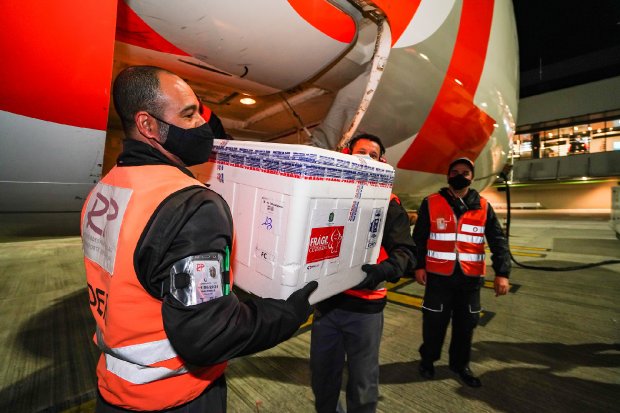 Trabalhadores de colete refletivo e máscara facial descarregam uma caixa térmica branca rotulada como “frágil” e com símbolos de material biológico, provavelmente contendo vacinas ou medicamentos, do compartimento de carga de um avião. A cena ocorre à noite, em uma pista de aeroporto, com o avião ainda estacionado e parcialmente visível ao fundo. A operação parece cuidadosa e coordenada, destacando a importância do conteúdo transportado.