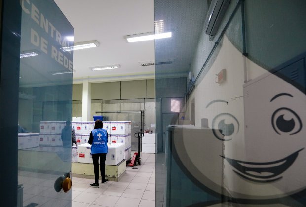  Sala de armazenamento refrigerado com caixas térmicas brancas empilhadas, identificadas com etiquetas de material biológico. Uma profissional da saúde, vestindo colete azul com o símbolo do SUS, está de pé observando as caixas. A imagem é vista por meio de uma porta de vidro, onde aparece refletido o personagem Zé Gotinha, símbolo das campanhas de vacinação no Brasil. Na porta está escrito parcialmente “CENTRO DE REDE”. A cena sugere a chegada e organização de vacinas ou insumos médicos em um centro de distribuição.