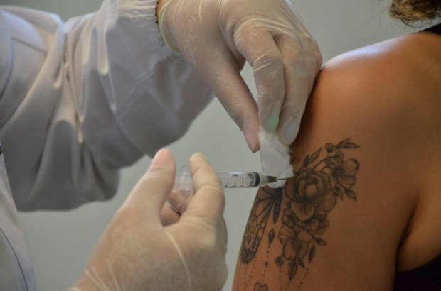 Mão com luva cirúrgica aplicando uma seringa em braço com tatuagem de flor no braço. 