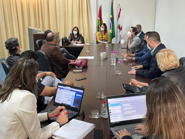 Reunião com 11 pessoas sentadas ao redor de uma mesa retangular em uma sala de reuniões. Três bandeiras estão posicionadas ao fundo: de Santa Catarina, do Brasil e de um órgão institucional. Algumas pessoas usam máscaras e outras não. Computadores, copos e blocos de anotações estão sobre a mesa.
