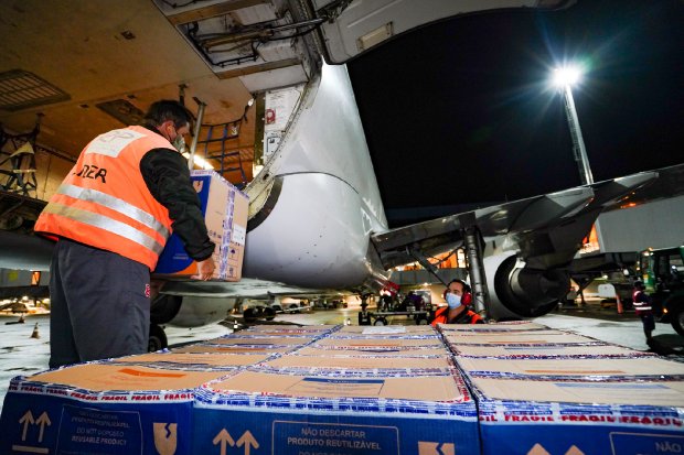 Homem de colete laranja e camisa preta retirando caixas de vacina do avião branco
