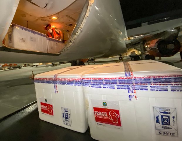 Funcionário descarrega caixas térmicas identificadas como contendo material frágil e sensível à temperatura de dentro do compartimento de carga de um avião. A cena ocorre à noite, em pista de aeroporto, e sugere o transporte de vacinas ou medicamentos.
