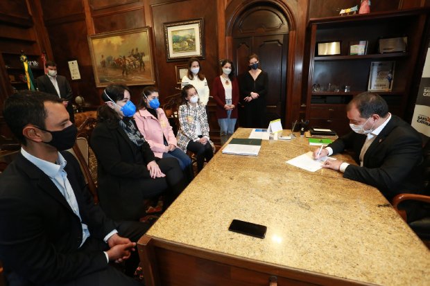 Reunião oficial com autoridades estaduais em gabinete. Ao centro, gestor assina documento acompanhado por equipe técnica e representantes de diversas instituições, todos usando máscaras de proteção.