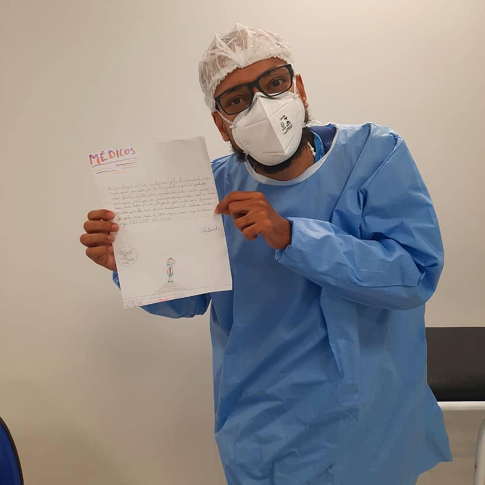 Médico segurando carta com desenho que recebeu de uma criança o desenho contém um personagem de máscara em cima de um semicírculo marrom. 
