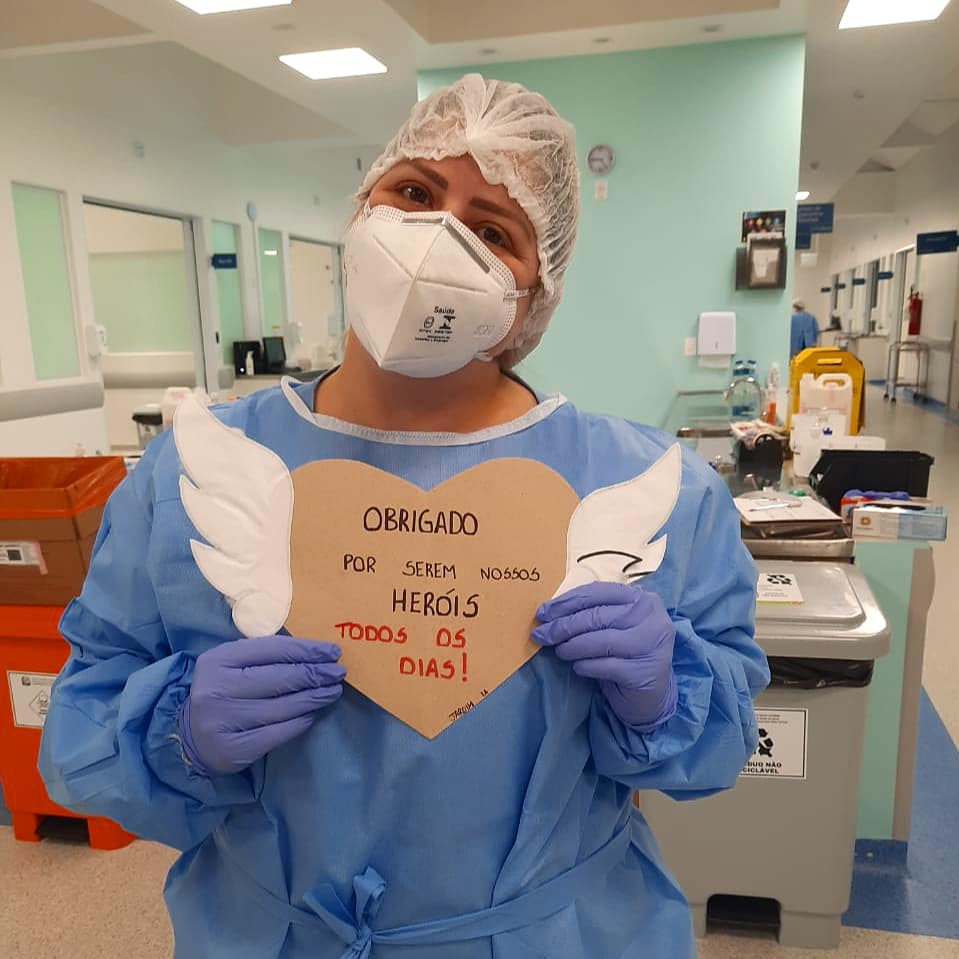 Médica segurando papelão recortado em formato de coração com asas com texto 'obrigado por serem nossos heróis todos os dias'