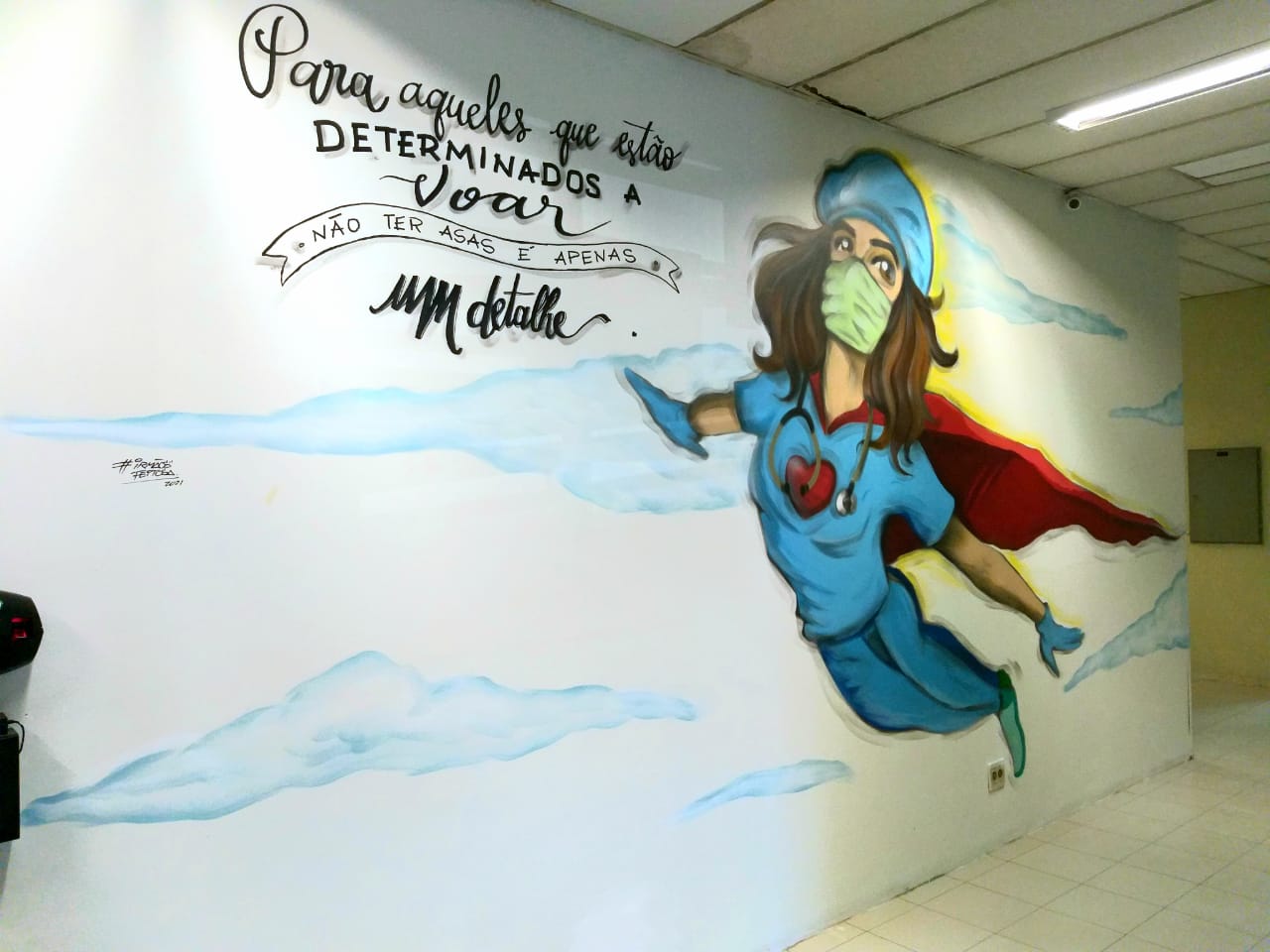 Mural pintado em parede interna de hospital retrata uma profissional de saúde vestida como super-heroína, com jaleco azul, estetoscópio no pescoço, capa vermelha e máscara verde, voando entre nuvens. Acima, a frase: Para aqueles que estão determinados a voar, não ter asas é apenas um detalhe. A arte é assinada por @sonhosdetinta.