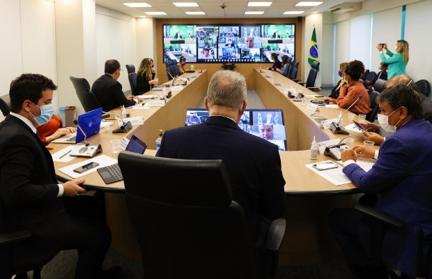 Mesa com diversas autoridades, no fundo um telão com uma videochamada sendo transmitida.