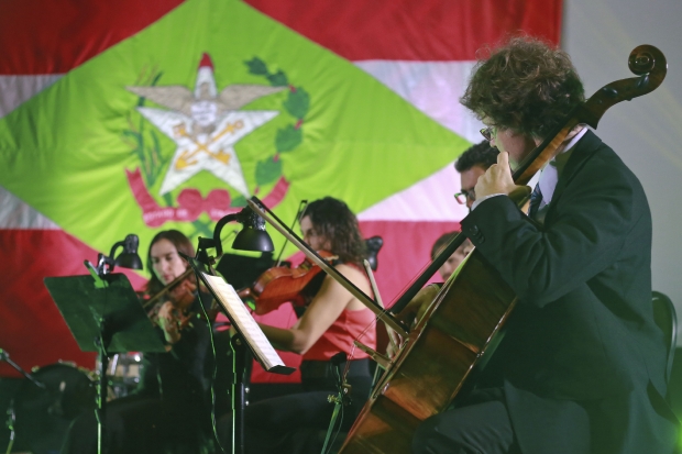 Orquestra com instrumentistas. Ao fundo, bandeira de Santa Catarina